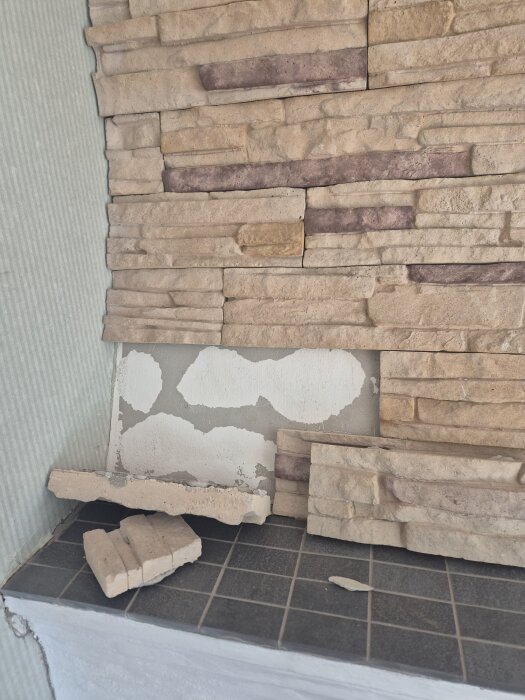 Lösa stenar på murstock med synlig vit puts bakom, några stenar har fallit ned på den svarta kakelbeklädda ytan.