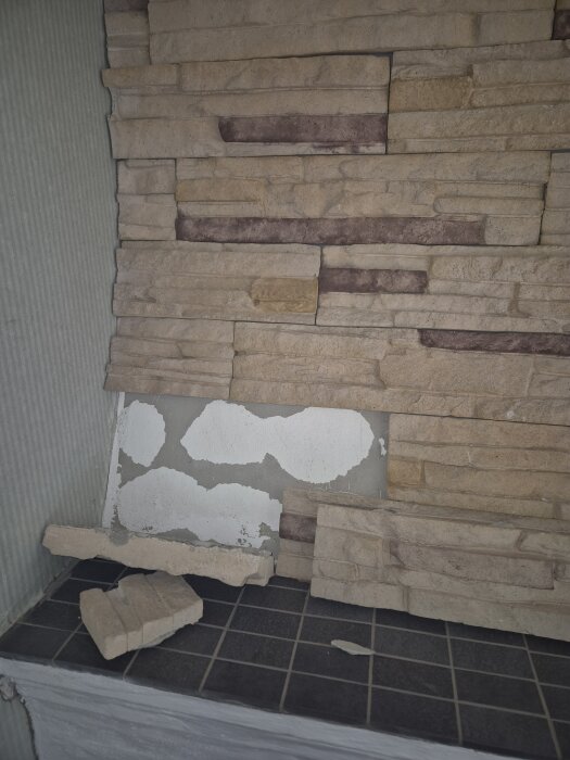 En murstock med lösa stenar där limmet torkat, visar vit puts bakom och några stenar som fallit ner på en svart kakelplattform.