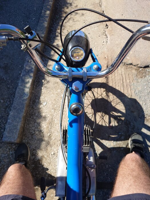 Vy från förarens perspektiv på en blå moped med styrstång och hastighetsmätare synlig, parkerad på en grusväg.