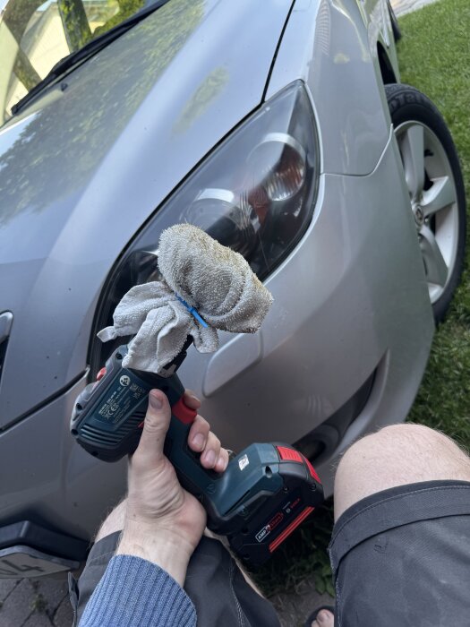 En person polerar en bilstrålkastare med en skruvdragare, borste och trasa fäst med buntband.