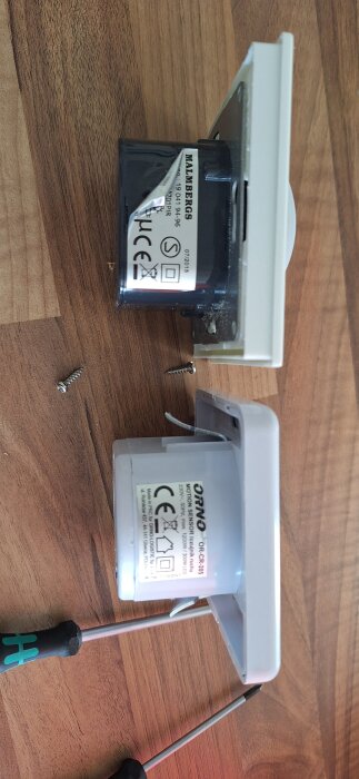 Två urkopplade rörelsedetektorer av märkena Malmbergs och Orno på ett träbord, bredvid två skruvmejslar och lösa skruvar.