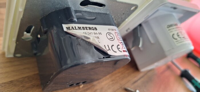 Närbild på en skadad rörelsevakt av märket Malmbergs, med en smält etikett, på ett träbord.