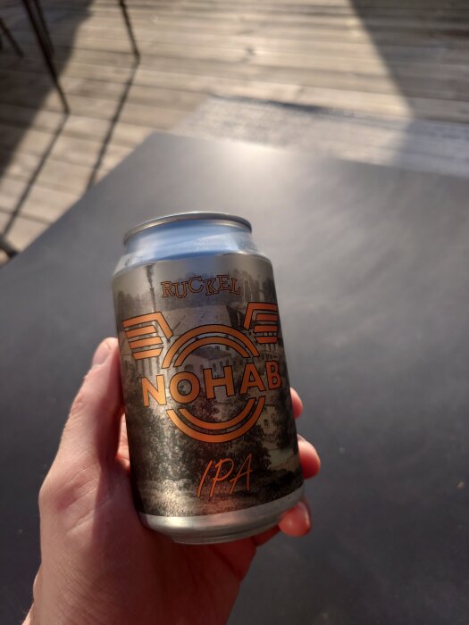 En hand håller en burk med etiketten "RUCKEL NOHAB IPA" mot en bakgrund av ett trädeck och ett bord.