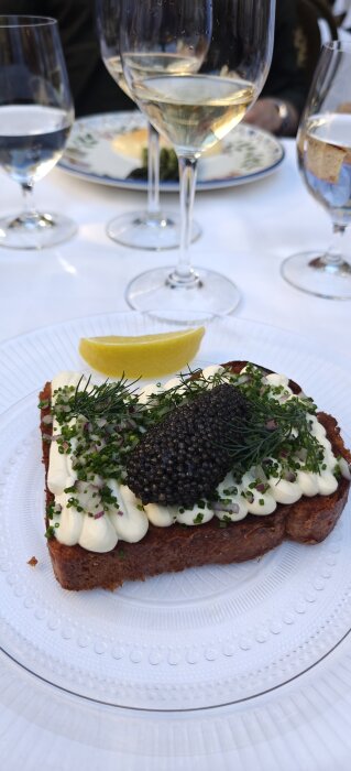 En tallrik med en smörgås toppad med kaviar, dill, gräslök och citron, omgiven av vinglas.