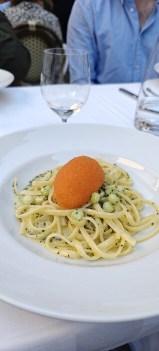 En vit tallrik med pasta dekorerad med gröna ärtor och en orange topping, placerad på en dukad bordsduk.