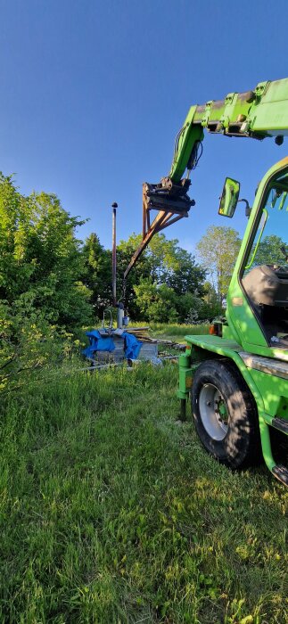 Grön maskin flyttar kamin i trädgård; blå skyddstäckning, träd i bakgrunden.