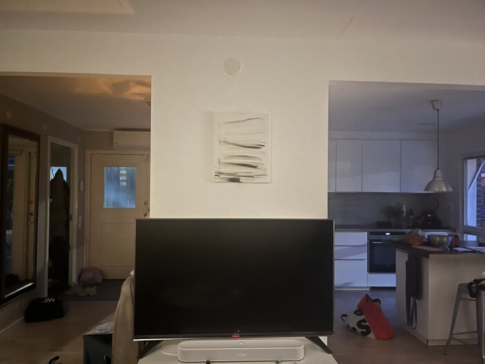 En modern inomhusmiljö med en vit vägg under en TV, ett kök till höger och en dörr till vänster.