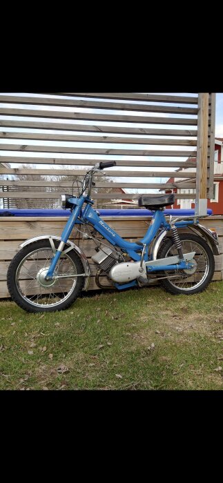 Blå moped av märket Mustang med originaldäck från Trelleborg, placerad mot en trävägg i en trädgård. Lackerad ram och välbevarad.
