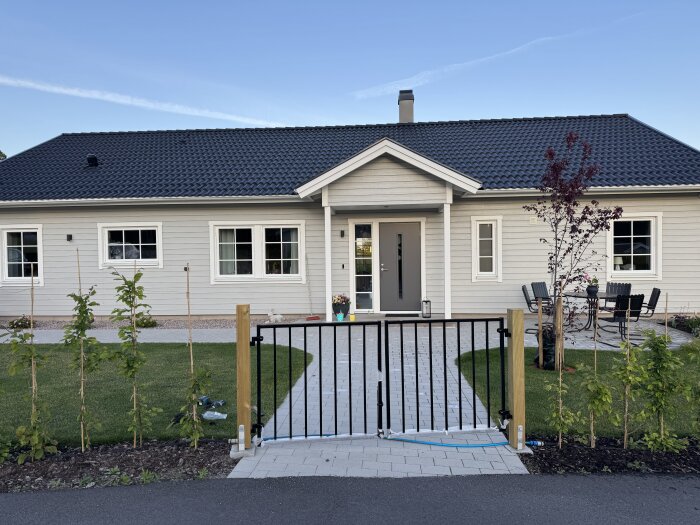 Svart dubbelgrind framför modernt ettplanshus med grå panel, vita fönsterkarmar och svart tak, omgiven av en stenlagd gång och trädgård.