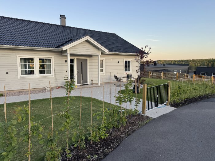 En ljusgrå villa med en grusad uppfart och två grindar i trä och metall som leder till en trädgård, omgivna av grönska och en asfalterad väg.