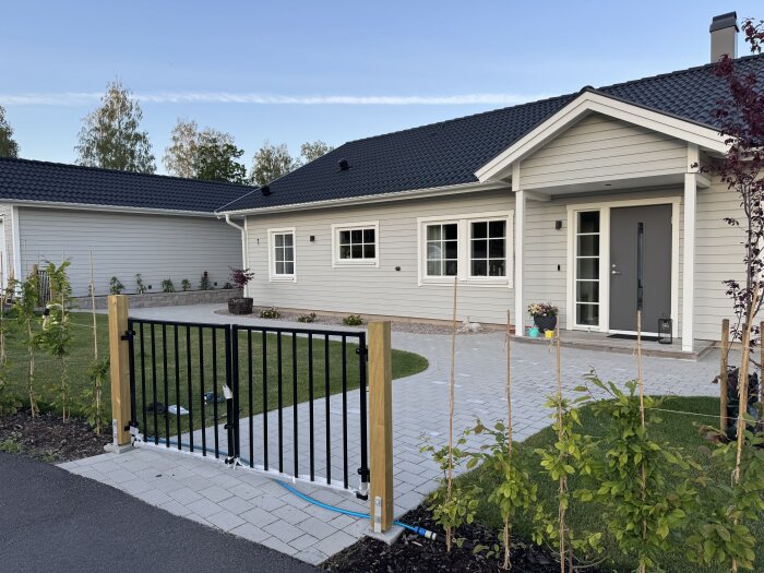 Dubbelgrind framför ett ljust hus med mörkt tak, asfalterad infart och grönska på sidorna. Garageuppfarten ligger till vänster från gatan sett.