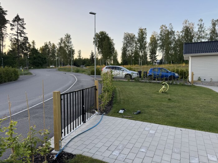 Stenlagd infart med gräsmatta och en svart grind vid sidan, träd och parkerade bilar syns i bakgrunden.