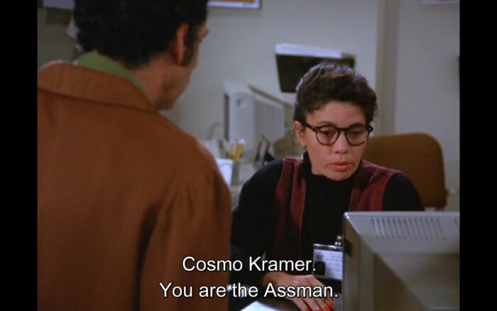 En person pratar med en kvinna vid ett skrivbord; texten säger "Cosmo Kramer. You are the Assman.