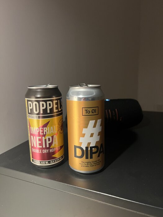 Två burkar öl på en svart yta, en från Poppels med etikett "Imperial NEIPA" och en från To Øl med etikett "#DIPA". JBL-högtalare i bakgrunden.
