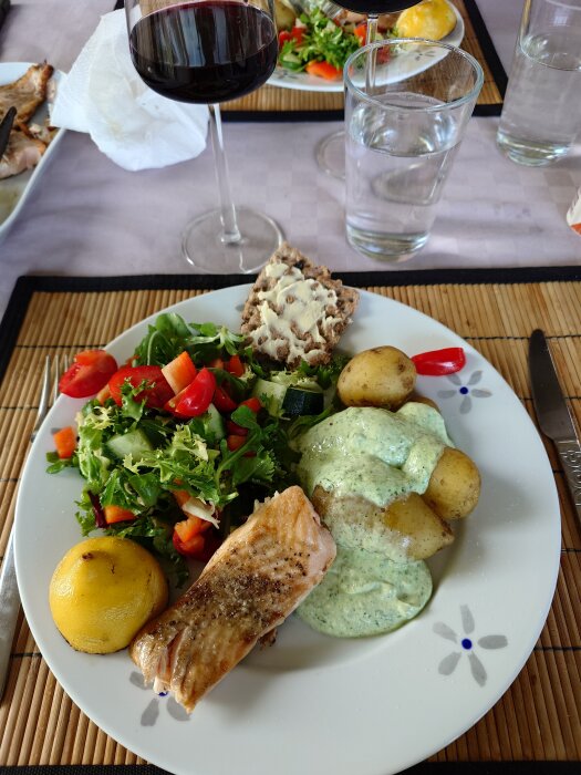 Tallrik med grillad lax, kokt potatis med basilikasås, grönsallad och brödskiva. Ett glas rött vin och vatten syns också på bordet.