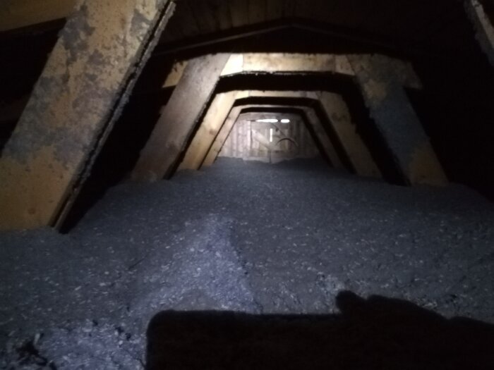 Vindsutrymme med tilläggsisolering av lösull, gamla träbjälkar och en yttervägg synlig i bakgrunden, svagt upplyst.