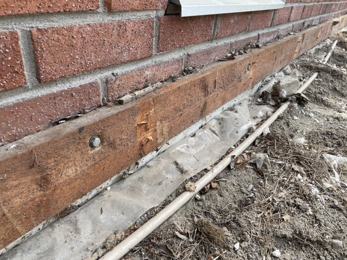 Träbalk fäst vid en tegelvägg ovanför plast och ojämn mark med rör och löv omkring.