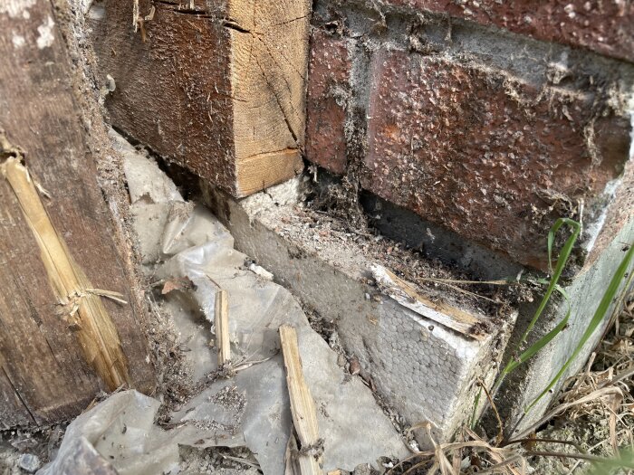 Närbild på en grund med träbjälkar och tegel, där plast och isoleringsmaterial syns i marknivå. Träet och plasten är slitna och dammiga.