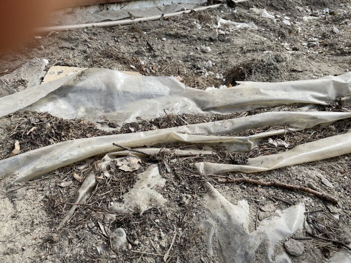 Plast över jord med ojämnheter, stenar och pinnar, omgiven av grus och torra löv, som en del av ett förberett byggområde för en altan.