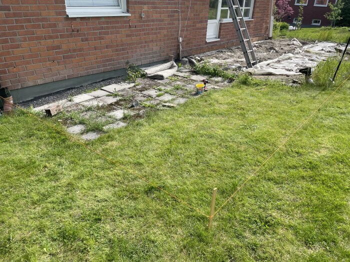 Gräsmatta med orange snöre markerar område, tegelhus i bakgrunden, stege lutad mot väggen, stenar och markskydd syns.