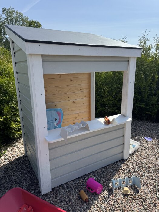 En liten grå och vit glasskiosk/lekstuga i trä med öppet fönster, omgiven av leksaker och grus, under en klarblå himmel.