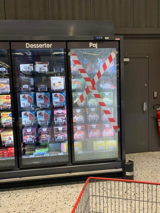 Trasig frys på stormarknad med tejpad ruta och varor som pajer och desserter synliga genom glaset. Kundvagn i förgrunden.