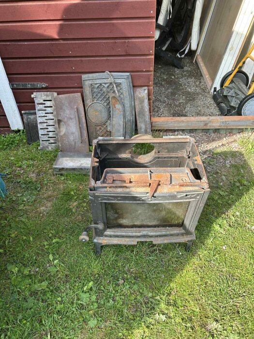 Rostig Jøtul vedeldad kamin och lösa delar framför ett förråd, grön gräsmatta och röd trävägg i bakgrunden.