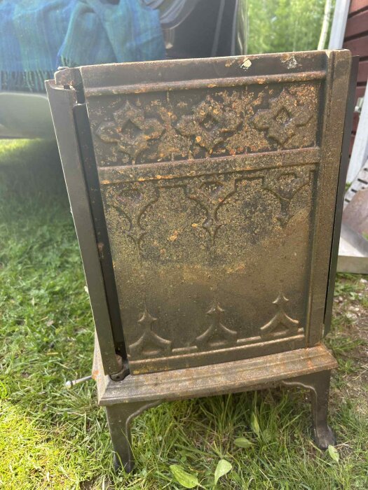 Rostig Jøtul vedeldad kamin (f100) stående utomhus i gräs, med utsirad dekor och ytor som visar tecken på rostspridning.