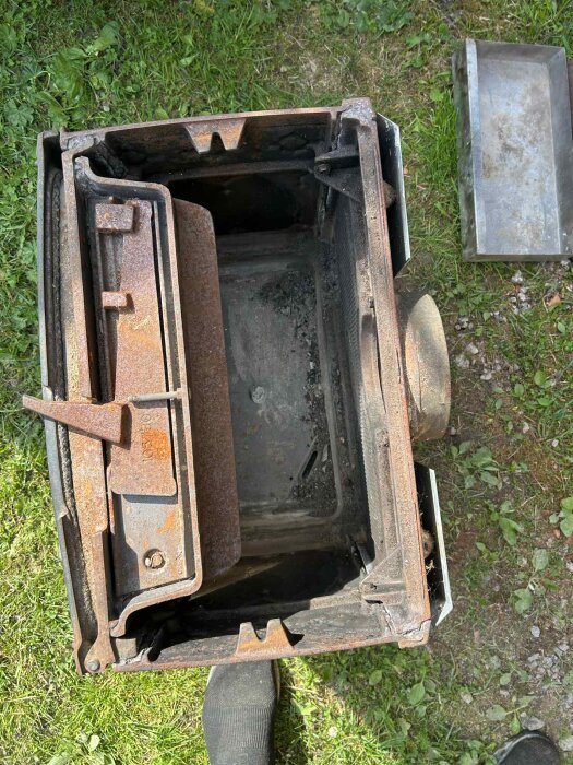 Rostig Jøtul vedeldad kamin, modell F100, sedd uppifrån med löstagna delar, placerad på en gräsmatta med en fot i bild.