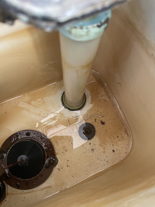 Invändig bild av en gammal toalettcistern från 60-talet som visar röranslutningar och ventiler, delvis fyllt med vatten och sediment.