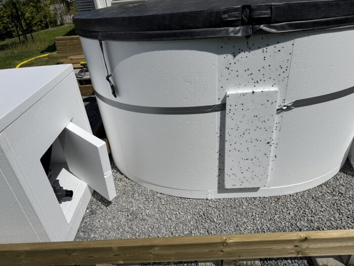 Isolerad rund terrasspool med ett vitt frigolitskydd och öppnad tekniklåda på grusunderlag, träregel synlig i förgrunden.