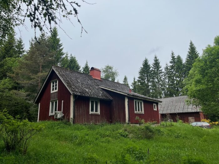 Rött torp med gamla, slitna eternitplattor på taket omgiven av grönska, i behov av renovering och byte av takmaterial.
