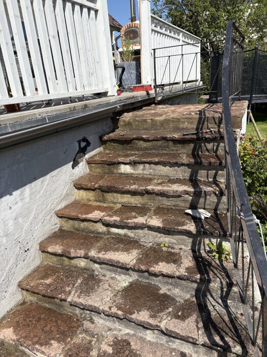 Utomhustrappa med ojämnt placerade och skiktade stenar, flagnande fogar och ett fall mot ett vitt räcke och svart järnspjälsverk på sidan.