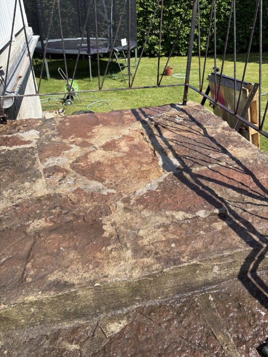 Ojämnt stenunderlag på en altantrappa efter avlägsnande av stenar, med synliga större stenar och skiftande tjocklekar. Trädgård i bakgrunden.