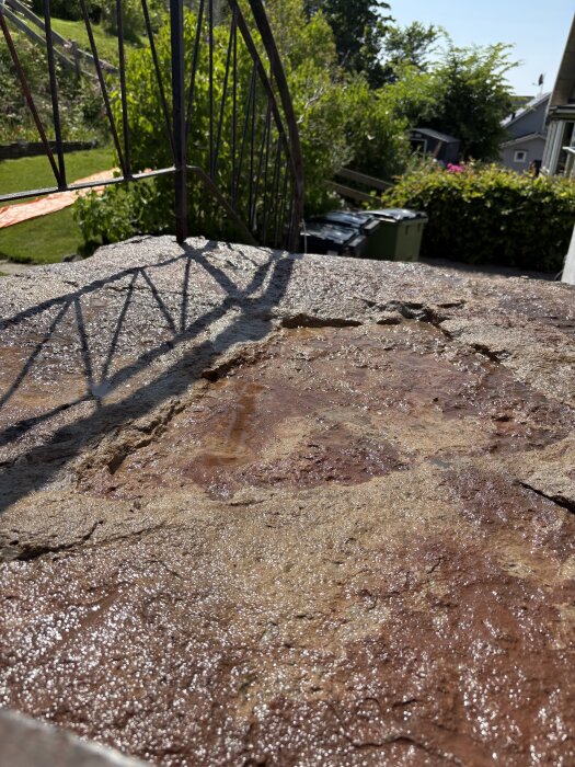 Ojämnt underlag av större stenar på övre platån av altantrappa efter borttagning och rengöring, med skugga från räcke, omgivande grönska.