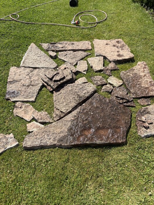 Stenar från en altantrappa ligger utspridda på en gräsmatta, varierande i storlek och tjocklek, med en trädgårdsslang i bakgrunden.