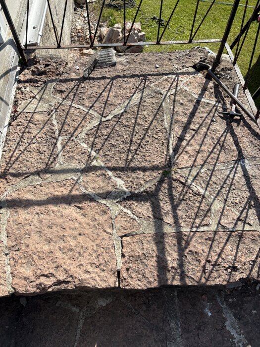 Ojämn stenyta på altantrappa med synliga fogar och skuggor från ett räcke. Trappan har delvis borttagna stenar och synliga färska sprickor.