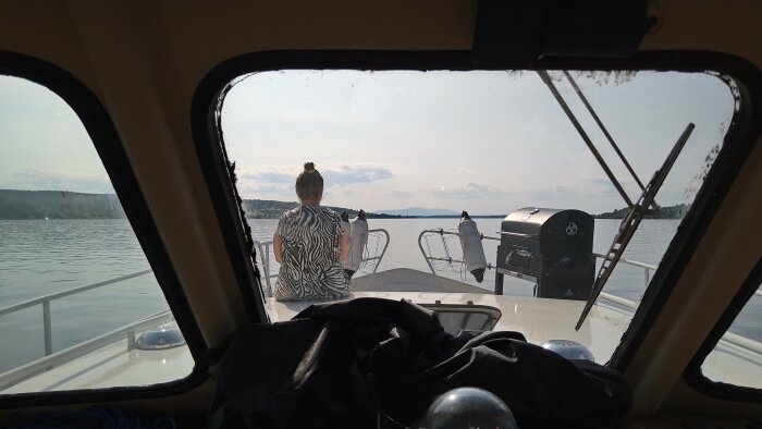 Utsikt från båtens cockpit, en person sitter i fören och blickar ut över vattnet med grill synlig till höger.