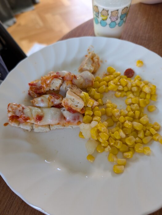 Tallrik med brödbitar täckta med smält ost och tomatsås, bredvid en hög av majs. Bakgrund med golv och ett glas med mjölk.