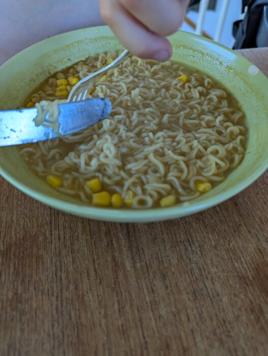 En skål med nudelsoppa och majs serveras, en hand håller en gaffel och kniv ovanför soppskålen.