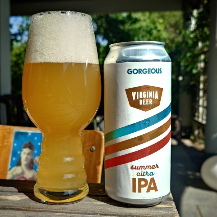 En kanna med gul IPA bredvid en ölburk märkt "Virginia Beer Summer Citra IPA" på ett bord i en solig trädgård.