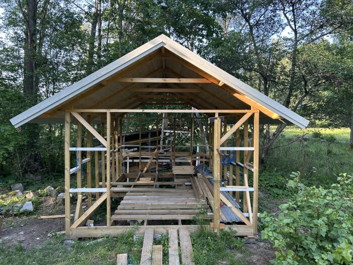 Byggprojekt av trästruktur, troligtvis en mindre byggnad under konstruktion i skogsmiljö, med synliga takstolar och öppna väggar.