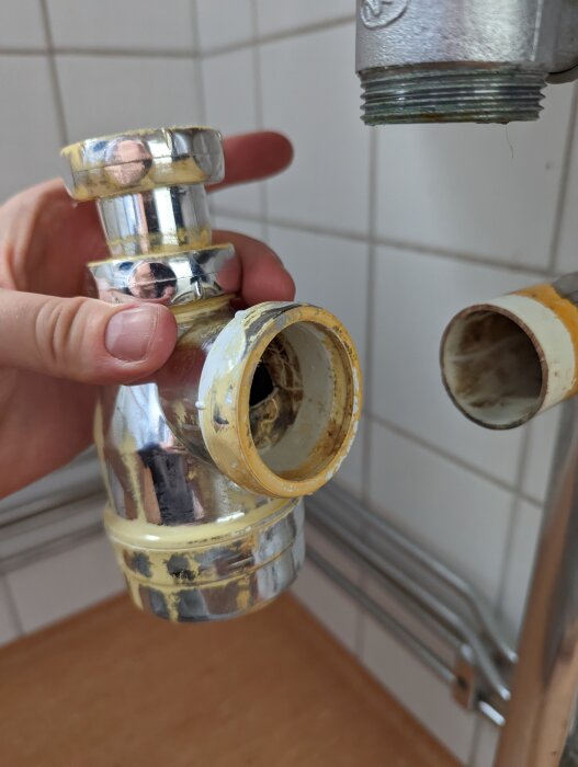 En hand håller ett avloppsrör och vattenlås under tvättställ i badrum, där anslutningen saknar tätning och orsakar läckage.