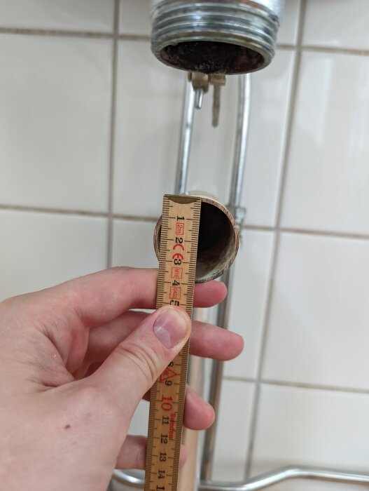 En hand håller en linjal mot änden av ett avloppsrör under ett tvättställ, där anslutningen verkar läcka.