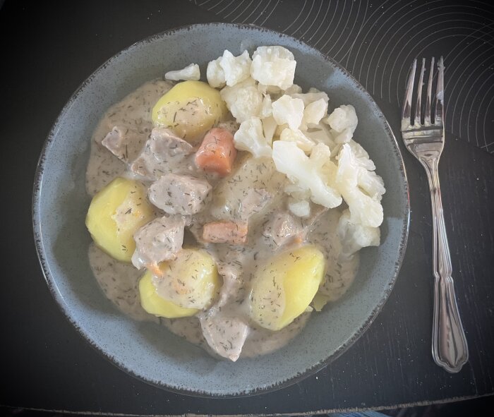 Tallrik med dillkött i dillsås, serverat med kokt potatis och blomkål. Fork bredvid tallriken.