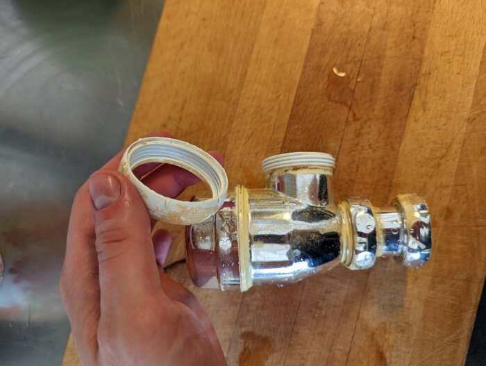 En hand håller en plastdel bredvid ett metalliskt vattenlås på en träyta.