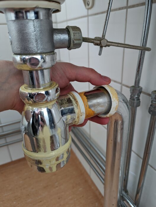 Användare håller i ett förkromat vattenlås under ett handfat i ett kök eller badrum, redo för byte eller reparation.