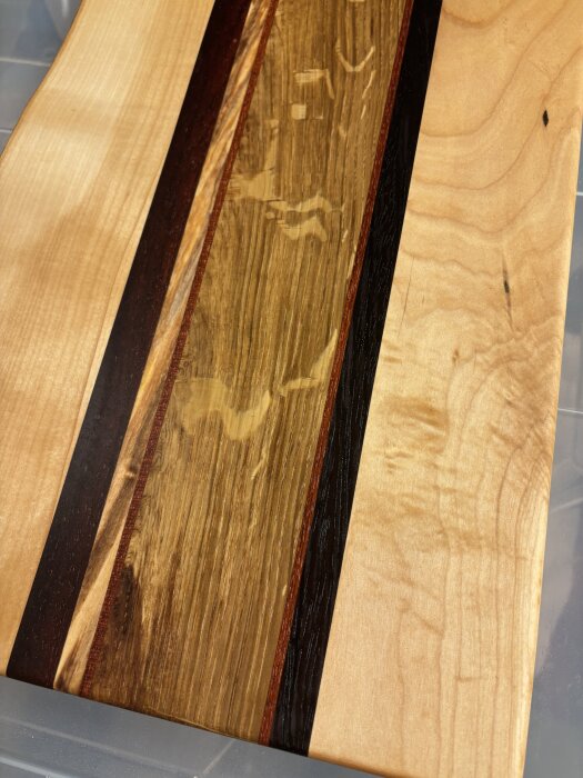 En handgjord skärbräda av flera träslag, inklusive lönn och valnöt, med mörka och ljusa partier samt råkant i svart bets för unik design.