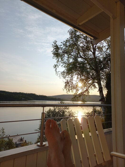 Solnedgång över sjö sedd från altan med träd och fötter vilandes på en stol.