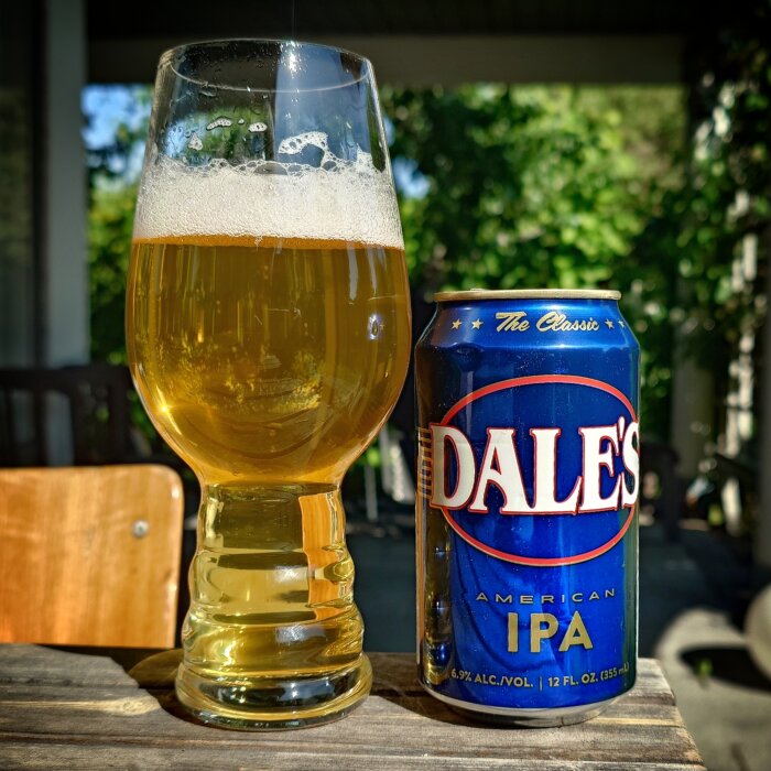 Blå burk med Dales American IPA och ett glas fyllt med öl på ett träbord utomhus.